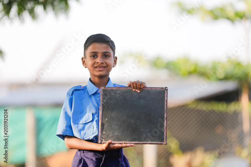happy indian villager schoolboy in uniform sits outdoors and holds up a slate