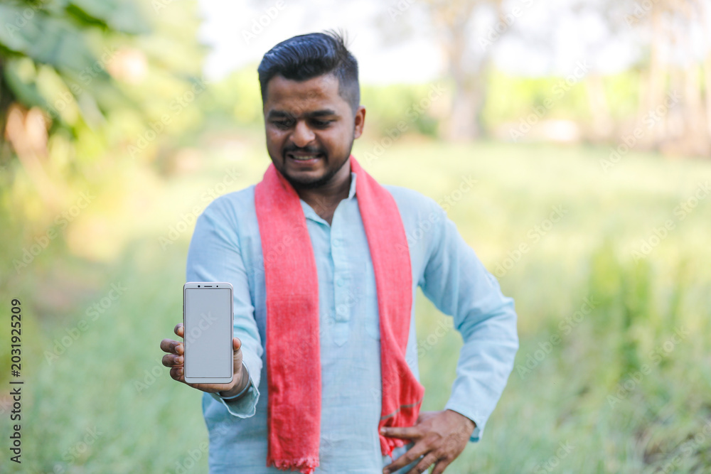 Fototapeta premium young indian farmer showing smartphone screen sitting at green agricultural field
