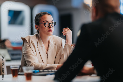 A professional businesswoman listens attentively to a colleague during a meeting. She participates in a focused office conversation with a business partner.