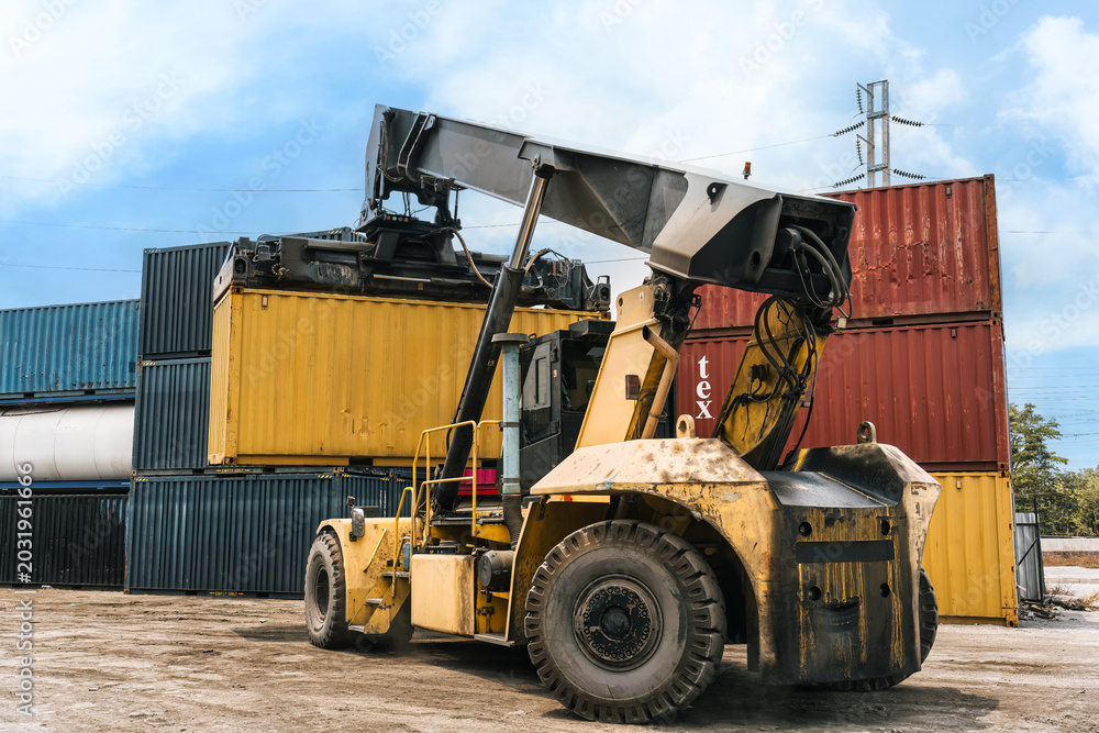 Obraz premium Loaded container handler forklift moving cargo container in crowded shipping yard at port terminal