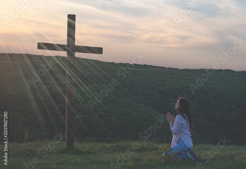 A young girl or woman kneels near a wooden crucifix and prays. Forgiveness of sins through atonement. Death of Jesus. Easter. Prayer.