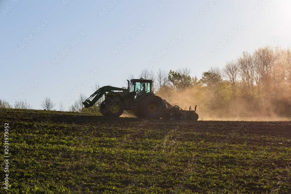 Fototapeta premium Backlit silhouette of unrecognizable man plowing field in heavy-duty agricultural machine raising dust, St-Augustin-de-Desmaures, Quebec, Canada