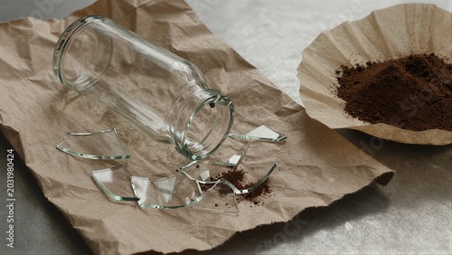 Broken glass jar spills coffee grounds on brown paper symbolizing