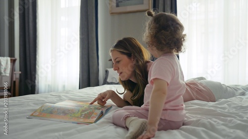 Mother and young daughter bonding over a children's picture book while comfortably lying on a bed at home, sharing a special moment of early learning and family connection