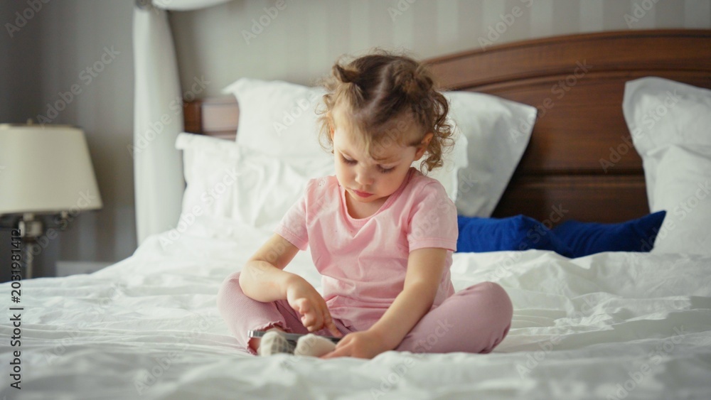 Fototapeta premium Toddler girl sitting cross legged on a comfortable bed, engaged with a mobile device, representing early childhood technology use, learning, and home based entertainment