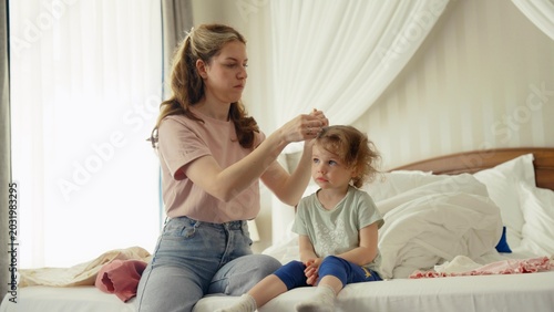 Tableau sur toile Mother tending to her little daughter's hair while sitting on the bed, creating