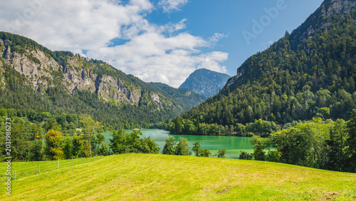 Canvas Print Landscape shows green grass field and lake surrounded by mountains under blue sk