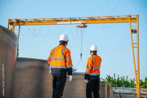 Professional engineer teamwork in safety uniform watching heavy steel metal material lifting by gantry crane at industrial factory outdoor site. Commercial construction business infrastructure project