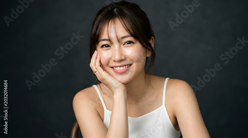 Asian Woman Smiling: A radiant young Asian woman with a gentle smile and her hand resting on her cheek, captured in a moment of serene happiness, looking directly at the viewer.