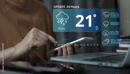 Fotografía Close-up of a person using a smartphone to check a digital weather forecast dashboard, showing a rainy and thunderstorm prediction at 21 degrees with a 24-hour update graph, next to a laptop