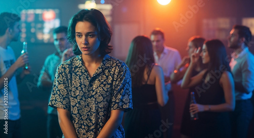 Shy person at party stands alone in crowded bar, looking reserved while others socialize. Background shows guests chatting, emphasizing feelings of social anxiety.