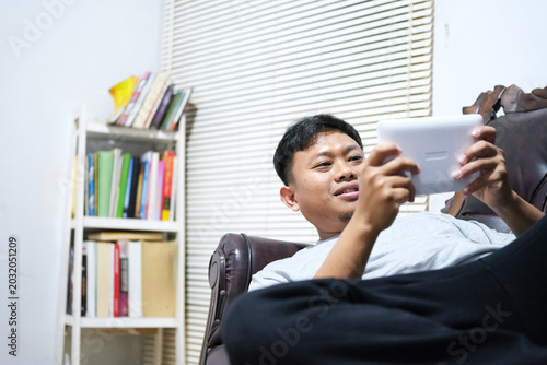 Bild auf Leinwand A cheerful Asian man is having fun playing video games on his smartphone while comfortably lying on a sofa