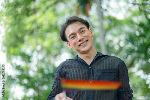 Strikingly healthy Asian man with short hair handsome and somewhat effeminate is waving pride flag representing his sexual diversity and LGBTQ identity. He is a man who is attracted to other men.

