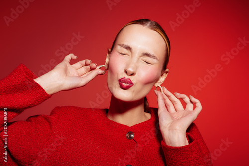 Photos Young woman in red jacket makes playful face with closed eyes and puckered lips on red background