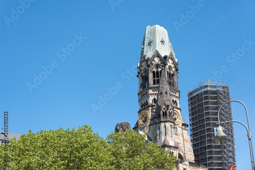 Kaiser Wilhelm Memorial Church rises above London plane trees in Berlin. Scaffolding and clear blue sky frame the historic tower.