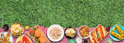 Outdoor summer food BBQ bottom border with hot dogs, hamburger and sides. Above view on a red and white check cloth and grass banner background.