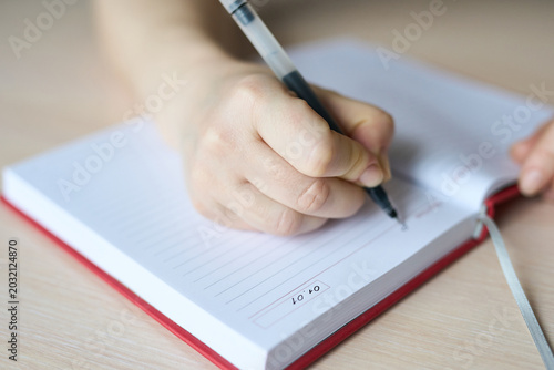 Child Hand Filling Diary Entry In Red Bound Notebook On Wooden Surface, Pen Poised Over Lined Page With Date Box, Indoor Daylight Close-Up Documenting Daily Notes And Planning Routine.