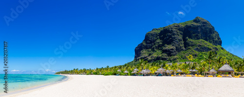 A luxurious white sand beach with palm trees, sun loungers, and umbrellas in Mauritius. A beautiful tropical beach with Le Morne Mountain as a backdrop. The coastline of an African island.