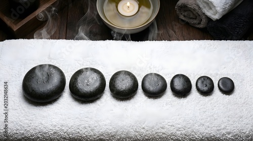 Row of graduated black spa stones with water droplets on a white towel, steaming bowl with a candle in the background, zen atmosphere for relaxation and wellness therapy sessions