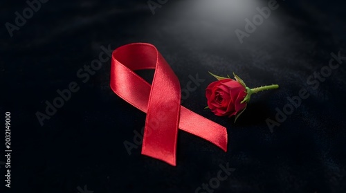 A single bright red satin awareness ribbon placed next to a small, fresh red rose blossom on a dark, soft-textured black fabric background, symbolizing support, love, and remembrance