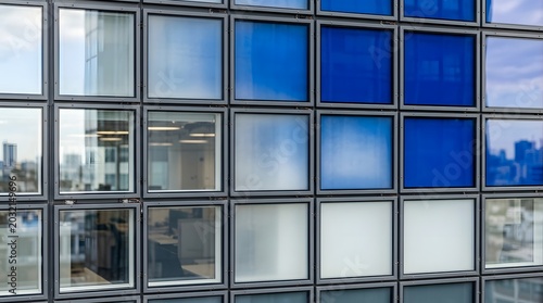 Exterior facade of a modern office building featuring a grid of square glass windows with varying shades of blue, white, and transparent reflections of the city skyline and interior