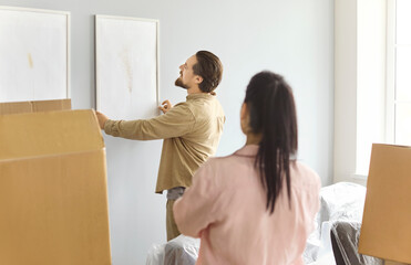 Couple moving decorating wall with frames apartment. Cardboard boxes and wrapped furniture signal relocation as they plan layout share teamwork and begin unpacking. Concept shows new home setup.