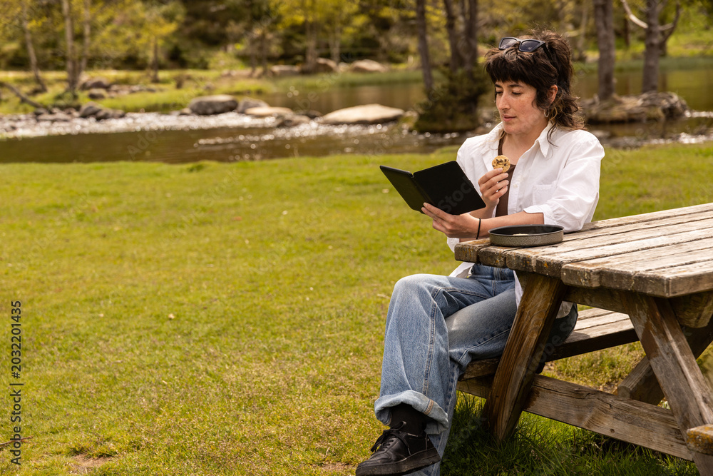 Obraz premium Woman sits at a wooden picnic table in a sunny park. She wears a white shirt and blue jeans. She reads a book while eating a snack. Her expression is calm and relaxed. Peaceful outdoor activity