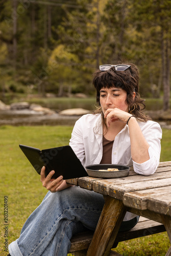 Woman sits at wooden picnic table in grassy forest. She holds tablet, eats from a bowl of food. Sunglasses rest on her head, wearing white shirt. Her focused expression suggests calm concentration