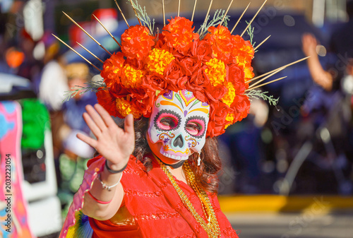Fotografi EL PASO, TEXAS – NOVEMBER 01, 2025: A festival participant wearing an elaborate orange and red marigold floral headdress and a colorful skull mask waves during annual Dia de los Muertos celebration