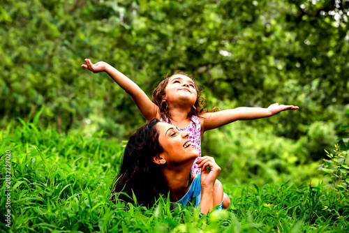 Joyful Mother and Child in Nature: A young girl with arms outstretched embraces the world, perched on her mother's back as they relax in a lush, green grassy field amidst a vibrant forest.