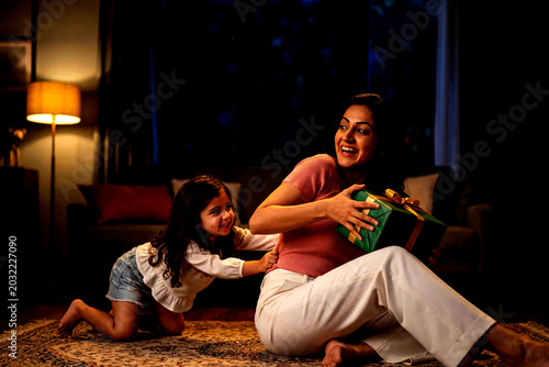 A Mother's Joyful Surprise: A young daughter playfully tugs at her mother's arm as she eagerly receives a beautifully wrapped gift, illuminated by soft ambient light in a cozy home setting.