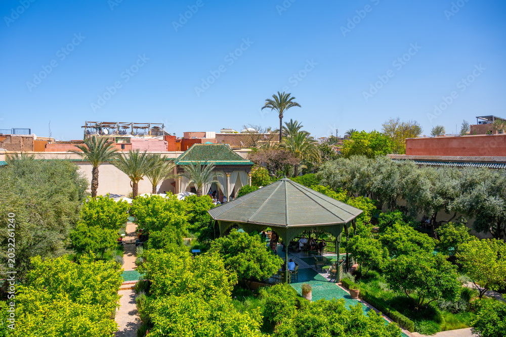 Fototapeta premium Visitors enjoy the beauty of Secret Garden in Marrakesh. The garden features palm trees, green plants, and pathways for people to stroll. It is a popular spot for relaxation and conversation.