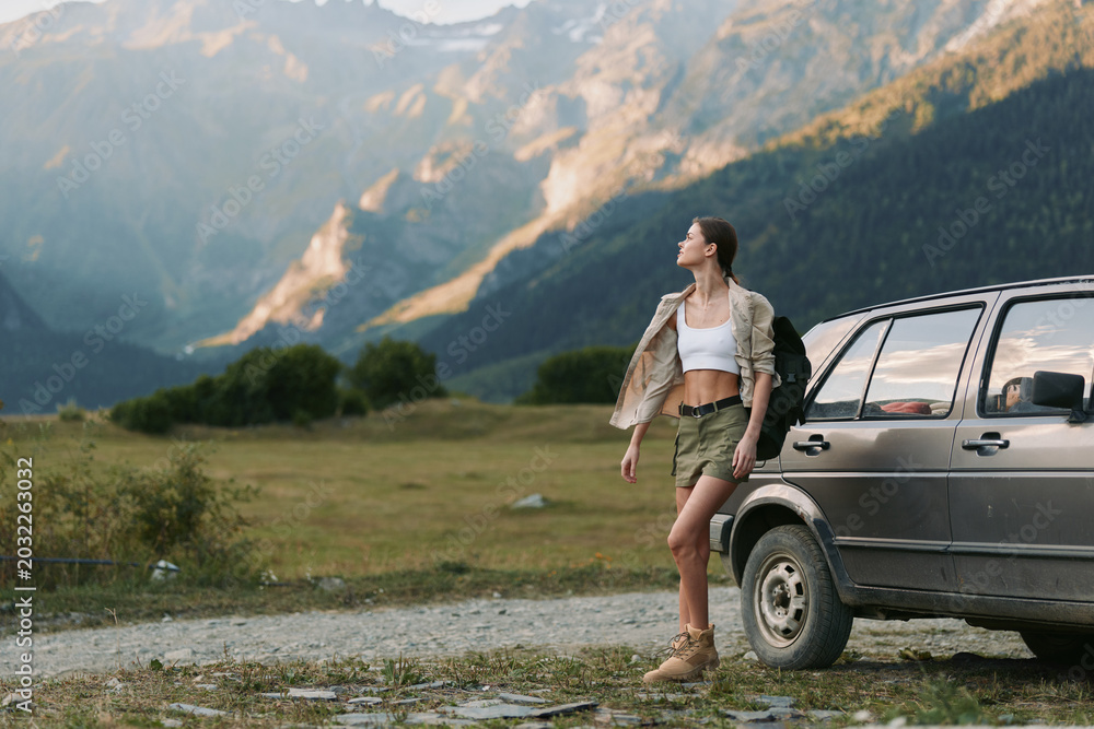 Fototapeta premium Woman stands beside a rugged car in a mountain meadow, wearing shorts and a cropped top with a jacket, embracing an adventurous outdoor lifestyle and wide horizon, wind in hair, sense of freedom,