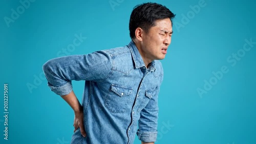 Asian Man Wearing Denim Shirt Suffering From Lower Back Pain Standing Against Blue Background With Harsh Lighting