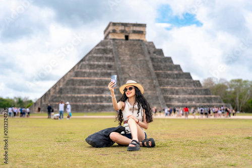 Young smiling Latina woman happily taking a selfie, expressing joy excitement while visiting Kukulkan pyramid at Chichen Itza, Yucatan Mexico, wearing summer outfit with hat, sunglasses in sunny day
