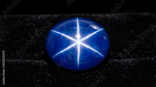 A polished blue star sapphire gemstone featuring a distinct six-rayed asterism light effect, resting on a dark black velvet surface, macro close-up view of a precious cabochon stone