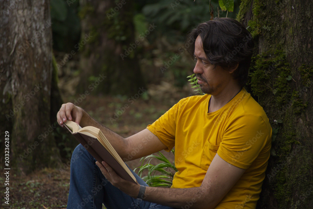 Obraz premium Side view of an adult man leaning against a tree trunk while reading a book outdoors.