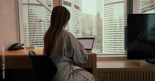 Woman in a robe typing on a laptop at a desk in a modern hotel room overlooking urban skyscrapers during the day. Slow zoom in camera movement from behind.