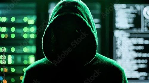 A person in a hoodie with green lighting looks at screens with code