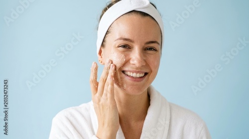 A young woman with fair skin is applying a facial scrub to her face. She is wearing a white headband and a white robe.