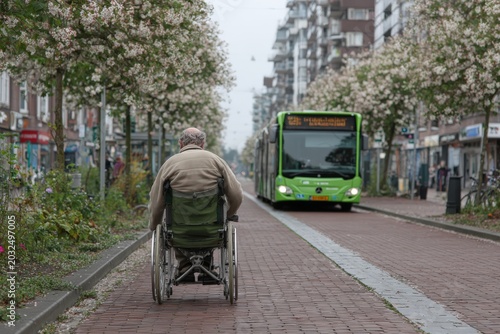 Elderly man in wheelchair on a brick pathway surrounded by flowering trees, with a green city bus approaching in a residential urban area