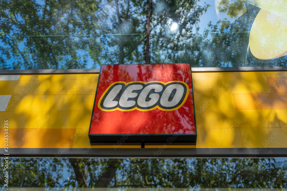 Obraz premium Germany, Berlin May 23, 2026 A LEGO store sign stands against a yellow facade and reflective glass. Reflections of London plane trees and blue sky add color to the urban storefront.