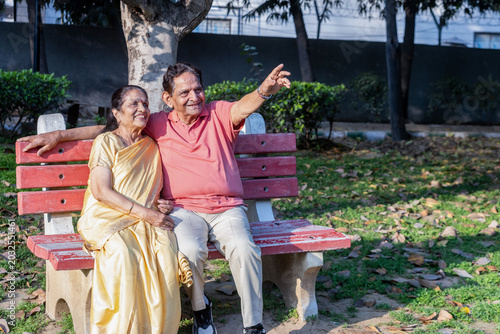 Indian elderly couple sitting on park bench, smiling and pointing forward, representing future goals and companionship.