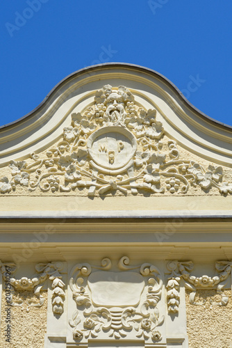 Obraz na plátně Low angle view of a historic house facade featuring a decorative arched tympanum with detailed plaster reliefs and a central crest under a clear blue sky