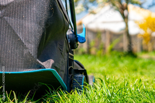 Low angle close up of a black and blue lawn mower on vibrant green grass in a sunny backyard garden, blurred wooden fence, trees, and house in the background with shallow depth of field.