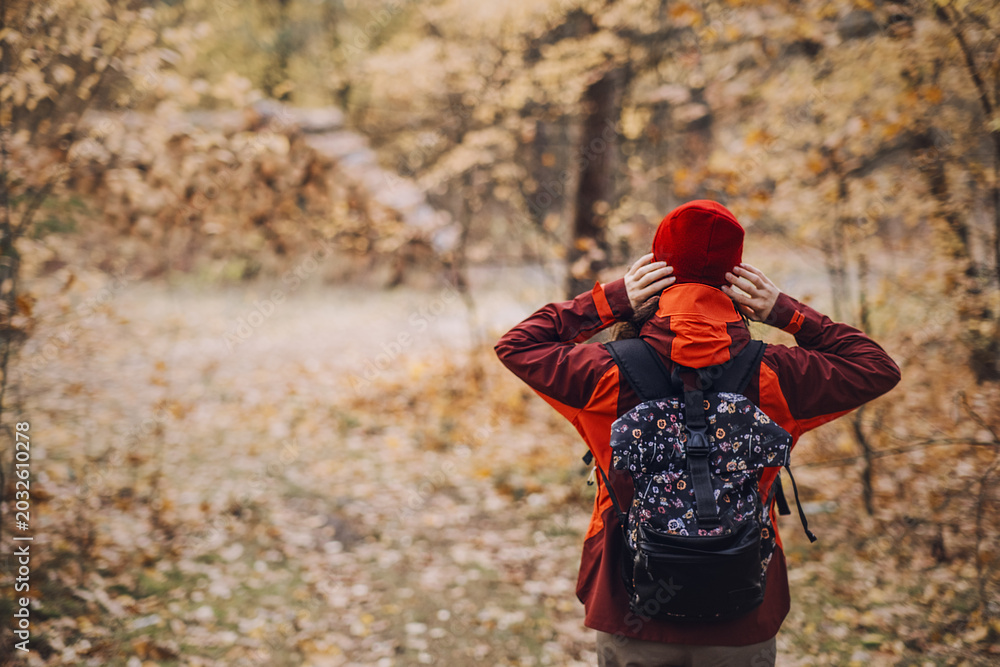 Naklejka premium Rear view of woman hiker adjusting hat while standing in autumn forest. nature-based therapy, mental resilience outdoors, forest bathing benefits.