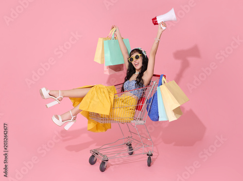 Shopping lifestyle asian woman sitting in cart with colorful bags and megaphone celebrating fashion sales joyful consumerism retail promotion trendy summer shopping and youthful spending culture.