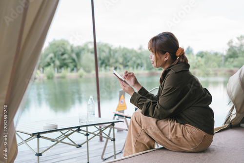 Digital communication and remote lifestyle concept young Asian woman using smartphone while relaxing on wooden porch of glamping tent by the lake during peaceful nature weekend getaway vacation