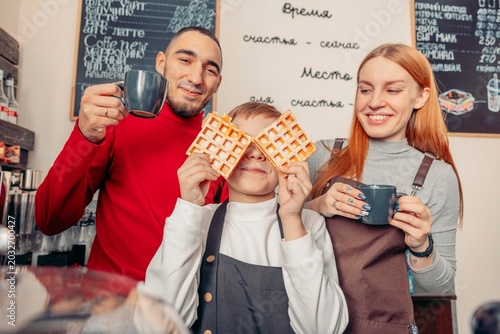 smiling parents and little boy having fun with coffee cups and fresh waffles in modern coffee shop interior. family lifestyle, breakfast and cafe culture