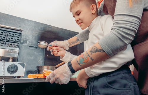 woman helping young boy sprinkle powdered sugar on freshly baked waffles in cafe kitchen. family bonding, cooking together and childhood learning concept